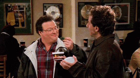 A Man In A Plaid Shirt Is Feeding Another Man Something GIF
