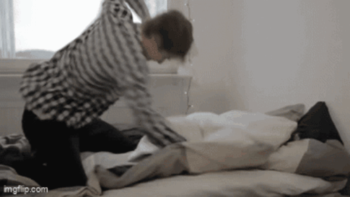 A Man In A Plaid Shirt Is Kneeling On A Bed With A Pillow . GIF