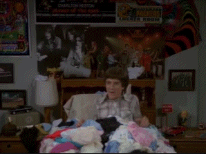 A Man In A Plaid Shirt Is Laying In A Bed With A Pile Of Clothes . GIF