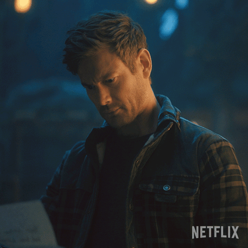 A Man In A Plaid Shirt Is Looking At A Piece Of Paper With Netflix Written On The Bottom GIF