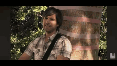 A Man In A Plaid Shirt Is Playing A Guitar In Front Of A Tree . GIF