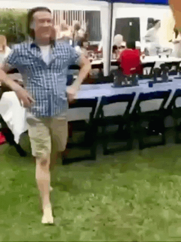 A Man In A Plaid Shirt Is Running Barefoot In The Grass In Front Of A Table And Chairs . GIF