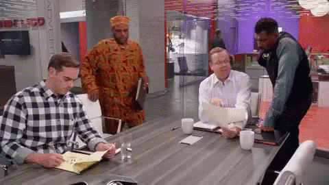 A Man In A Plaid Shirt Is Sitting At A Conference Table With Other Men GIF