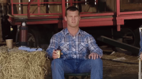 A Man In A Plaid Shirt Is Sitting In Front Of A Bale Of Hay GIF