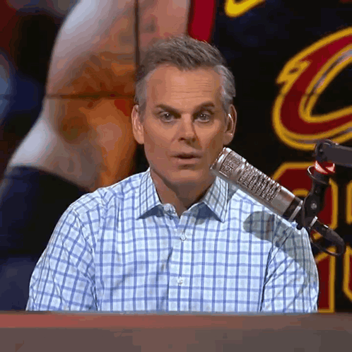 A Man In A Plaid Shirt Is Sitting In Front Of A Microphone With A Cleveland Cavaliers Jersey In The Background GIF