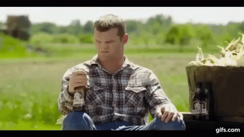 A Man In A Plaid Shirt Is Sitting On A Truck In A Field Holding A Glass Of Beer . GIF