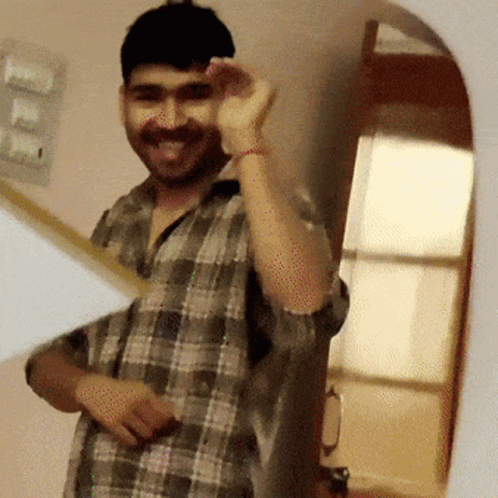 A Man In A Plaid Shirt Is Smiling And Waving At The Camera GIF