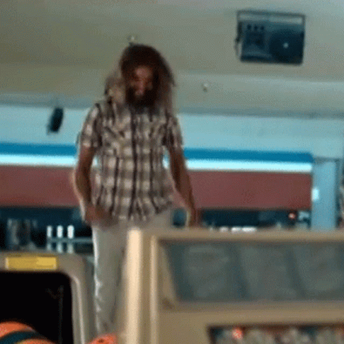 A Man In A Plaid Shirt Is Standing In A Bowling Alley Looking At A Bowling Ball GIF