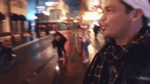 A Man In A Plaid Shirt Is Standing On A Wet City Street At Night . GIF