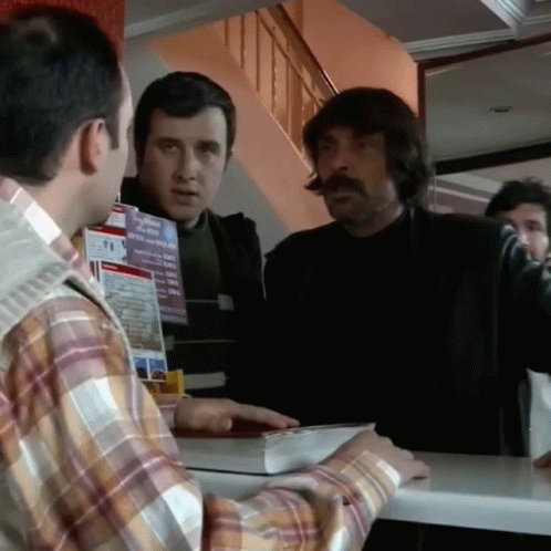 A Man In A Plaid Shirt Is Talking To Another Man In Front Of A Sign That Says T.i. GIF
