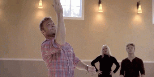 A Man In A Plaid Shirt Is Throwing A Frisbee In The Air While A Group Of People Are Watching . GIF