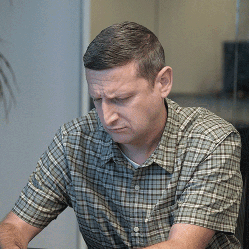 A Man In A Plaid Shirt Looks Down With A Serious Look On His Face GIF