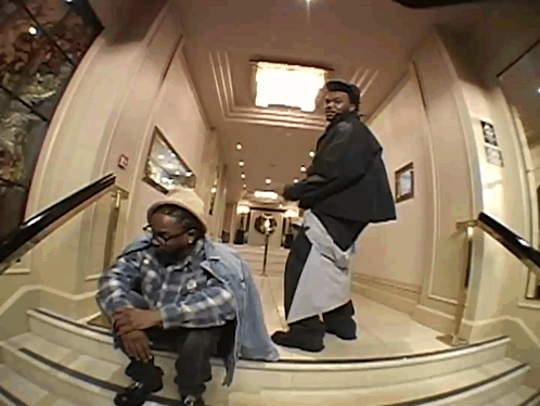 A Man In A Plaid Shirt Sits On A Set Of Stairs Next To Another Man In A Black Jacket GIF