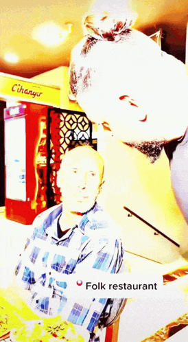 A Man In A Plaid Shirt Stands In Front Of A Sign That Says Folk Restaurant GIF