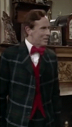 A Man In A Plaid Suit And Red Bow Tie Stands In Front Of A Fireplace GIF