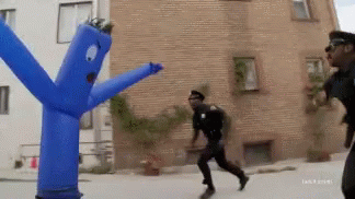 A Man In A Police Uniform Is Running Away From A Blue Inflatable Man With Arms And Legs GIF