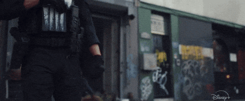 A Man In A Police Uniform Is Walking Down A Street In Front Of A Building With Graffiti On It . GIF