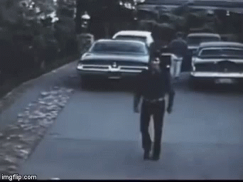 A Man In A Police Uniform Is Walking Down A Street In Front Of A Row Of Cars . GIF