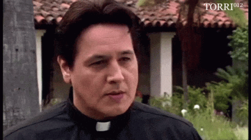 A Man In A Priest 'S Collar Is Standing In Front Of A Building With Torri912 Written In The Corner GIF