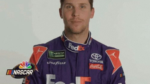 A Man In A Purple And Red Racing Suit Is Standing In Front Of A Nascar Sign . GIF