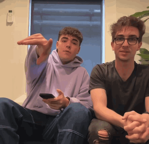 A Man In A Purple Hoodie Is Sitting Next To A Man In A Black Shirt GIF