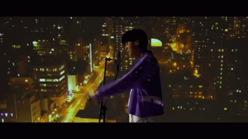 A Man In A Purple Jacket Sings Into A Microphone In Front Of A City At Night GIF