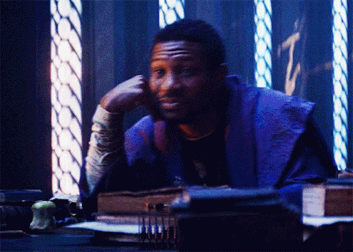 A Man In A Purple Robe Is Sitting At A Desk With His Hand On His Chin GIF