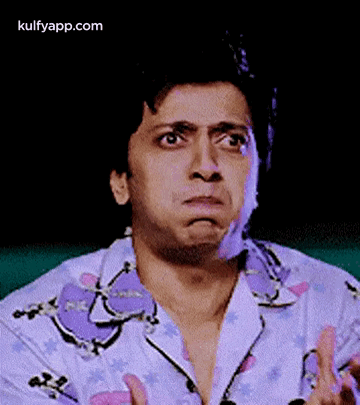 A Man In A Purple Shirt Is Making A Funny Face With His Hands . GIF