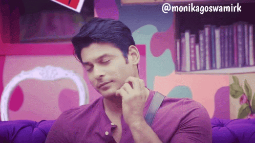 A Man In A Purple Shirt Is Sitting On A Purple Couch In Front Of A Bookshelf GIF