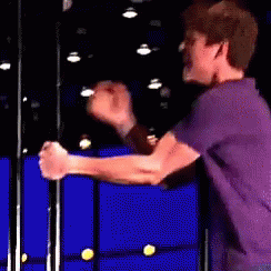 A Man In A Purple Shirt Is Standing In Front Of A Blue Wall With His Hands In The Air . GIF