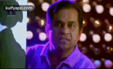 A Man In A Purple Shirt Is Standing In Front Of A Blurry Background Of Lights . GIF