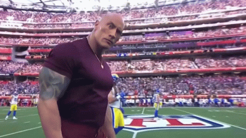 A Man In A Purple Shirt Is Standing On A Football Field In Front Of A Stadium Full Of People . GIF