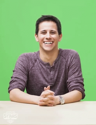 A Man In A Purple Shirt Sits At A Table With His Hands Folded In Front Of A Green Screen GIF