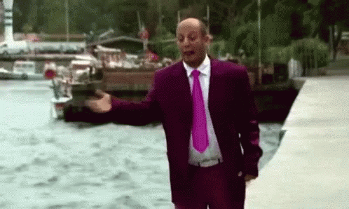A Man In A Purple Suit And Pink Tie Is Standing Next To A Body Of Water With His Arms Outstretched . GIF