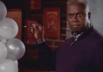 A Man In A Purple Sweater Is Standing In Front Of Balloons And Holding A Pen In His Hand . GIF