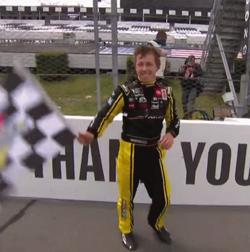 A Man In A Race Suit Holds A Flag In Front Of A Sign That Says Thank You GIF