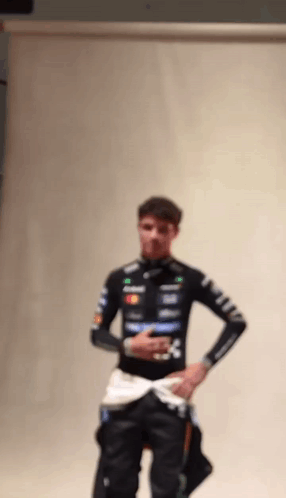 A Man In A Racing Suit Is Standing In Front Of A White Wall With His Hands In His Pockets . GIF