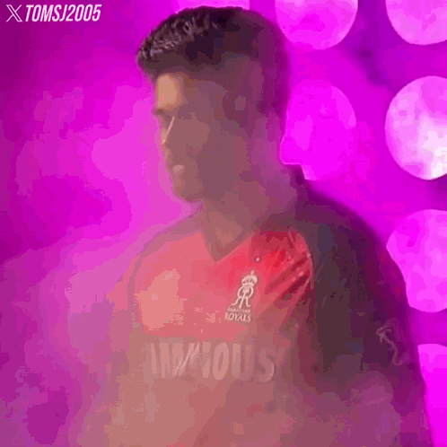 a man in a red and black cricket jersey is standing in front of a pink background .
