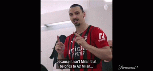 a man in a red and black jersey is standing in a locker room and talking .