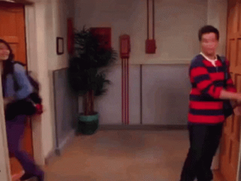 A Man In A Red And Blue Striped Shirt Is Opening A Door While A Woman Looks On . GIF