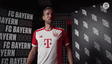 A Man In A Red And White Fc Bayern Jersey Stands In Front Of A Sign That Says Fc Bayern 1900 GIF