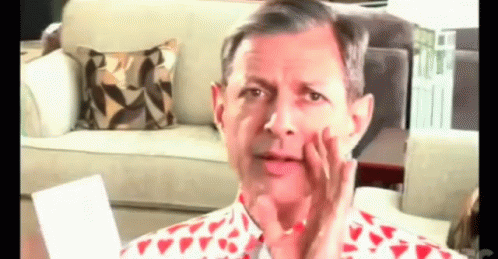A Man In A Red And White Shirt Is Sitting In Front Of A Couch Making A Funny Face . GIF