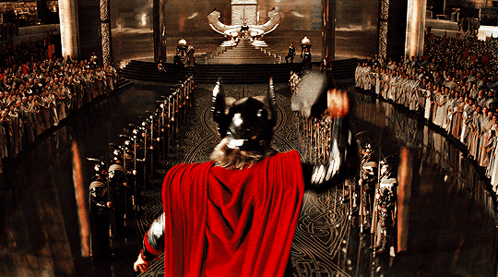 A Man In A Red Cape Is Holding A Hammer In Front Of A Large Crowd GIF