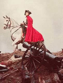 A Man In A Red Cape Is Riding A White Horse Next To A Cannon GIF