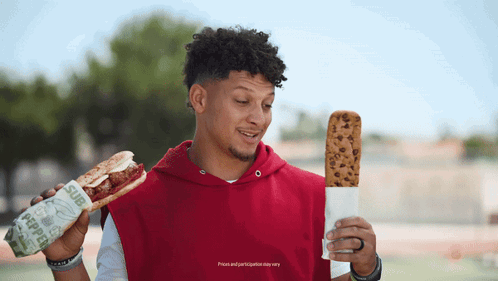 A Man In A Red Hoodie Is Holding A Sub Sandwich And A Cookie GIF