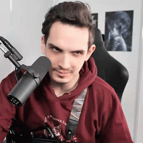 A Man In A Red Hoodie Is Sitting In Front Of A Microphone With A Picture Of A Skull On The Wall Behind Him GIF