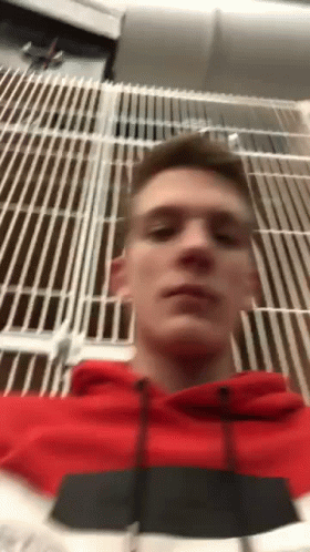 A Man In A Red Hoodie Is Taking A Selfie In Front Of A Metal Fence . GIF