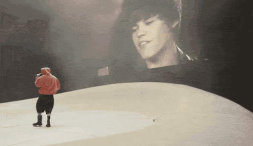 A Man In A Red Hoodie Stands In Front Of A Large Screen With A Picture Of Justin Bieber GIF
