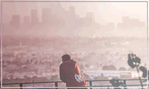 A Man In A Red Hoodie Stands On A Balcony With A City In The Background GIF