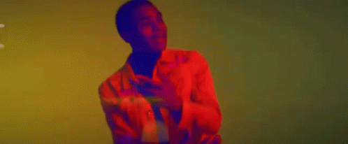 A Man In A Red Jacket And Black Turtleneck Is Dancing In Front Of A Yellow Wall . GIF